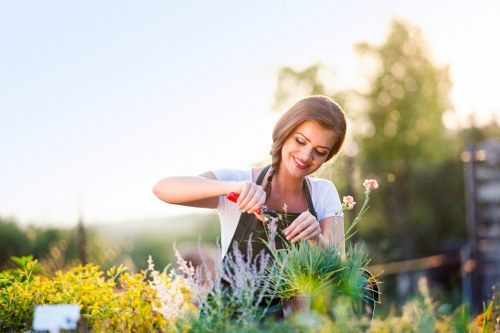 A young woman with a bright smile is tending to a lush garden bed in an outdoor space situated within a residential area near Fitzrovia. She is using a gardening trowel to carefully plant or tend to various green and flowering plants, including purple lavender-like foliage and pink blossoms. The garden features a well-maintained grass lawn with dense, vibrant green blades in the foreground, bordered by a variety of flower beds edged with soil and mulch. Behind her, mature trees and shrubs provide a natural backdrop, and a wooden fence runs along the side, indicating a private garden setting. The scene is illuminated by warm, natural sunlight, suggesting a clear, mild day in late spring or summer, with the garden appearing healthy and lively. This outdoor space showcases typical landscaping elements such as flower borders, lawn areas, and natural plantings, aligning with professional gardening and landscaping services offered by companies like Landscapers Fitzrovia that focus on elevating residential gardens in the local area, including postcode W1 in central London.