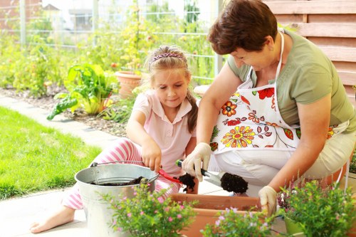 A woman and young girl are working together on a gardening project in a suburban backyard during daytime. The woman, wearing a floral apron and light green top, is kneeling on the grass and using gardening gloves to tend to a rectangular flower bed made of wooden sleepers, which contains various blooming plants with green leaves and colorful flowers. The girl, dressed in a pink and white striped outfit, is sitting on the grass beside her, assisting with planting or watering. The background shows a well-maintained lawn, a lush hedge, and a garden with flowering shrubs, potted plants, and a wire fence. The outdoor space appears bright with natural sunlight, suggesting fair weather. The scene emphasizes outdoor garden maintenance, including planting, flower bed care, and general landscaping, consistent with services offered by firms like Landscapers Fitzrovia in the local area near London or NW postcode regions relevant to Fitzrovia.