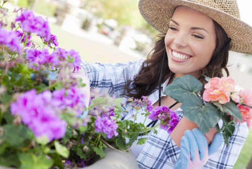 A woman with long dark hair, wearing a straw hat and blue gloves, is smiling as she tends to a colourful garden. She is in a landscaped outdoor space with vibrant purple and pink flowering plants, including petunias, which are arranged in containers or flower beds. The garden features a well-maintained grassy lawn area and a paved pathway visible in the background, suggesting a neat and organized layout suitable for outdoor gardening and maintenance services. The environment appears to be a bright, sunny day with natural light illuminating the scene, highlighting the lush green foliage and the vivid colours of the flowers, consistent with UK garden styles. This setting exemplifies typical elements that gardening specialists like Landscapers Fitzrovia may work with, including flower arrangements, plant care, and outdoor aesthetic enhancement in a private garden or similar landscaped area in London or nearby locales.