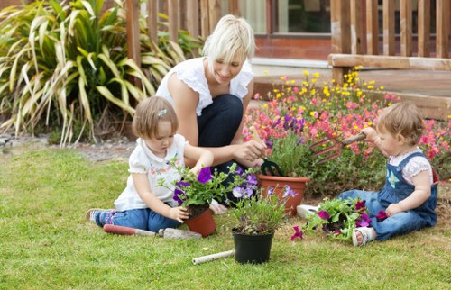 A woman with short blonde hair and wearing a white blouse is sitting on a well-maintained green lawn in a residential garden in Fitzrovia, engaging with two young children, a girl and a boy, who are seated on the grass. The children are dressed in casual clothing, with the girl in a floral top and the boy in a blue t-shirt and jeans. They are planting and tending to small potted garden plants, including colorful flowering plants and foliage. In the background, there are various garden elements such as a wooden deck with a railing, a brick pathway, and large ornamental grasses adding texture to the landscaped outdoor space. The garden is shown in natural daylight with bright weather, highlighting the vibrant colors of the flowers and the lush green grass. The scene captures a moment of outdoor leisure and gardening activity, reflecting typical services provided by Landscapers Fitzrovia, focused on garden maintenance and plant care within an urban outdoor environment in London postcode areas.