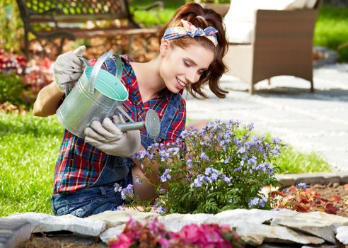 A young woman with a cheerful expression is kneeling in a well-maintained garden in Fitzrovia, tending to a colorful flower bed bordered by natural stone edging. She is wearing a plaid shirt, gardening gloves, and a headband, and is holding a small metal watering can in one hand while carefully watering a cluster of purple and pink blooming flowers. Behind her, a lush green lawn extends towards a wooden garden bench and a shaded patio area, possibly with outdoor furniture, under a clear sky. The garden features a mix of neatly trimmed shrubs, flowering plants, and small trees that add to the vibrant and inviting outdoor space. The soil in the flower bed appears rich and well-tended, with a combination of flowering plants and foliage providing natural variety. The scene captures an outdoor gardening activity during daylight hours, with natural sunlight highlighting the textures of leaves, flowers, and garden surfaces, illustrating the outdoor care and landscaping services offered by Landscapers Fitzrovia in the area of W1 postcode.