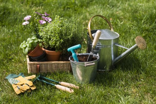 A neatly maintained garden scene featuring a lush green lawn with healthy, evenly cut grass in the foreground. In the background, there are potted plants with green foliage and purple flowers arranged on the ground. Nearby, there are gardening tools including a pair of yellow gardening gloves, small hand trowels with wooden handles, and a pair of long-handled pruning shears. A metal watering can with a wooden handle is positioned beside a silver bucket containing a small gardening trowel, all situated on the grass. The outdoor environment is well-lit with natural daylight, creating a bright and inviting atmosphere typical of a residential garden in Fitzrovia, London. The scene reflects general garden maintenance activities and outdoor care, aligning with landscaping and gardening services such as those offered by Landscapers Fitzrovia. The visible elements and their arrangement demonstrate an organized and thriving garden space suitable for ongoing landscaping and lawn care efforts.