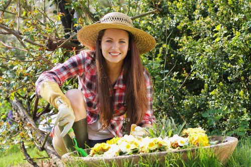 A young woman with long brown hair wearing a straw sun hat, red plaid shirt, and gardening gloves is kneeling in a lush, green garden. She is smiling at the camera while tending to a large, decorative wood or ceramic container filled with yellow and purple flowering plants. The garden features a well-maintained lawn with dense shrubbery and trees in the background, suggesting a vibrant outdoor space in Fitzrovia or nearby London. The scene is illuminated by natural daylight, with bright sunlight casting soft shadows across the grass and plants, indicating a clear, pleasant day. This image reflects gardening activity typical of residential or professional garden care, highlighting plant arrangement, garden maintenance, and outdoor landscaping work, aligning with services offered by landscapers fitting in central London areas. The garden's layout includes a neatly trimmed lawn, a variety of foliage, and a focus on flower display, supporting the context of expert gardening and landscaping services performed in urban outdoor environments.
