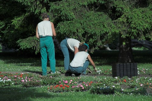 The image depicts a well-maintained garden area with a lush green lawn and a border of flowering plants in shades of pink, red, and white. In the background, there is a dense backdrop of mature trees with thick foliage, providing shade and natural privacy. Three individuals are actively engaged in garden maintenance; one person in teal trousers and a sleeveless top stands upright, while another in a white shirt and jeans is kneeling and tending to the flower bed, and a third, also kneeling, appears to be planting or weeding among the flowers. The garden features a combination of grass, flower beds, and a paved or paved-like surface area. The outdoor environment is lit by natural daylight, suggesting a clear or partly cloudy weather condition, suitable for gardening activities. These details reflect a typical landscaped outdoor space that a professional gardening or landscaping service such as Landscapers Fitzrovia might undertake in the Fitzrovia area, aligning with lawn care, planting, and garden maintenance services available locally.