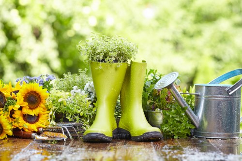 A pair of bright green rubber gardening boots filled with small white flowering plants are positioned on a wooden outdoor table. The table also holds a variety of garden-related items, including a bouquet of yellow sunflowers and purple flowers, a pair of pruning shears, and a garden watering can with a long spout. The background features a lush, green garden with dense foliage and sunlight filtering through the trees, creating a vibrant and fresh outdoor scene. The arrangement of gardening tools, plants, and decorative floral elements suggests preparation for planting or ongoing garden maintenance in a residential outdoor space within Fitzrovia or the surrounding London area, reflecting professional gardening practices by Landscapers Fitzrovia focusing on outdoor plant care and garden aesthetics.