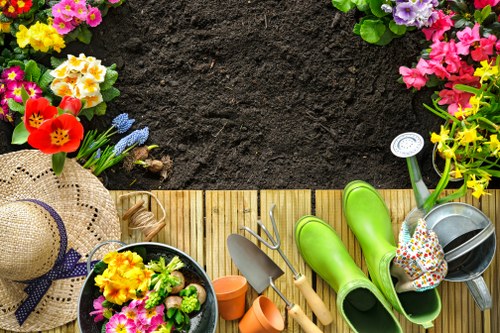 The image depicts a section of a garden in Fitzrovia featuring a wooden decking area bordered by a lush flower bed with vibrant pink, purple, yellow, and red blooms. In the foreground, there are garden tools and accessories, including a small trowel, a fork, a watering can, and a pair of bright green rubber gardening boots placed on the decking. A straw sun hat with a purple ribbon rests nearby, alongside potted plants with colorful flowers. The background shows freshly turned dark soil, ready for planting, with some planting pots and a garden fork lying on the wooden surface. The outdoor environment appears well-maintained and suitable for gardening activities, with natural daylight illuminating the scene. This setting, typical of a landscaped urban garden, highlights features that are common in professional gardening and landscape maintenance services in the Fitzrovia area, connecting details of plant care, garden organization, and outdoor aesthetic appeal managed by local landscapers like Landscapers Fitzzrovia.