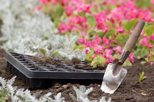 The image shows a close-up view of a garden bed with freshly turned soil, bordered by white flowering plants and small pink flowering plants in the background. A black seed tray with soil remnants is placed on the ground, indicating recent planting activity. A small garden trowel with a wooden handle rests partially in the soil, ready for use. The outdoor setting appears well-maintained, with natural daylight illuminating the scene, typical of spring or early summer in Fitzrovia. The arrangement suggests ongoing garden landscaping or planting work, which is within the scope of services offered by Landscapers Fitzzrovia. The scene highlights typical elements of garden preparation and planting, with a focus on soil, planting tools, and flower borders, reflecting the company's expertise in gardening and outdoor maintenance within central London.