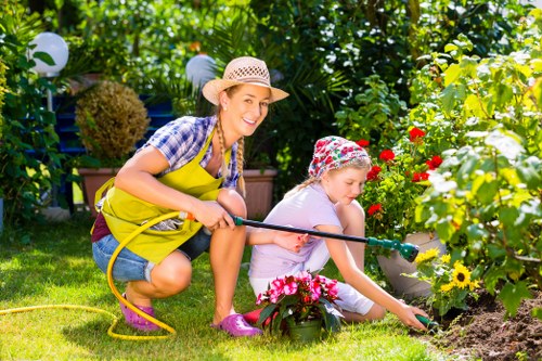 A woman and a young girl are gardening in a lush, well-maintained backyard, which features a vibrant flower bed with red, yellow, and pink blooms, bordered by a low raised wooden edge. The woman, wearing a straw hat, bright yellow apron, and casual clothing, is smiling and holding a garden watering can, while the girl, dressed in a floral hat and light-colored clothing, is kneeling on the grass, carefully planting or tending to the flowers with her hand. The garden area includes a neatly trimmed lawn with dense green foliage, trees, and shrubs in the background, and white outdoor lanterns hanging among the greenery. Sunlight filters through the leaves, creating a warm and inviting outdoor environment suitable for gardening activities, indicative of professional gardening and landscaping services in Fitzrovia or nearby areas, with a focus on outdoor garden maintenance and flower care.