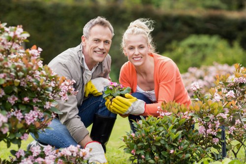 A smiling middle-aged man and woman with light skin are kneeling in a well-maintained garden, surrounded by blooming pink and purple flowering bushes. The man has short, light brown hair and is wearing a light grey shirt, while the woman has blonde hair tied back in a ponytail and is dressed in an orange sweater. They are both wearing yellow gardening gloves and holding a potted plant, indicating they are engaged in gardening activity. The garden features lush green grass in the foreground, with neatly trimmed hedges and a backdrop of trees and shrubs under natural daylight with slight overcast conditions, creating a vibrant yet calm outdoor environment typical of a high-quality garden maintained by professional landscapers in Fitzrovia.